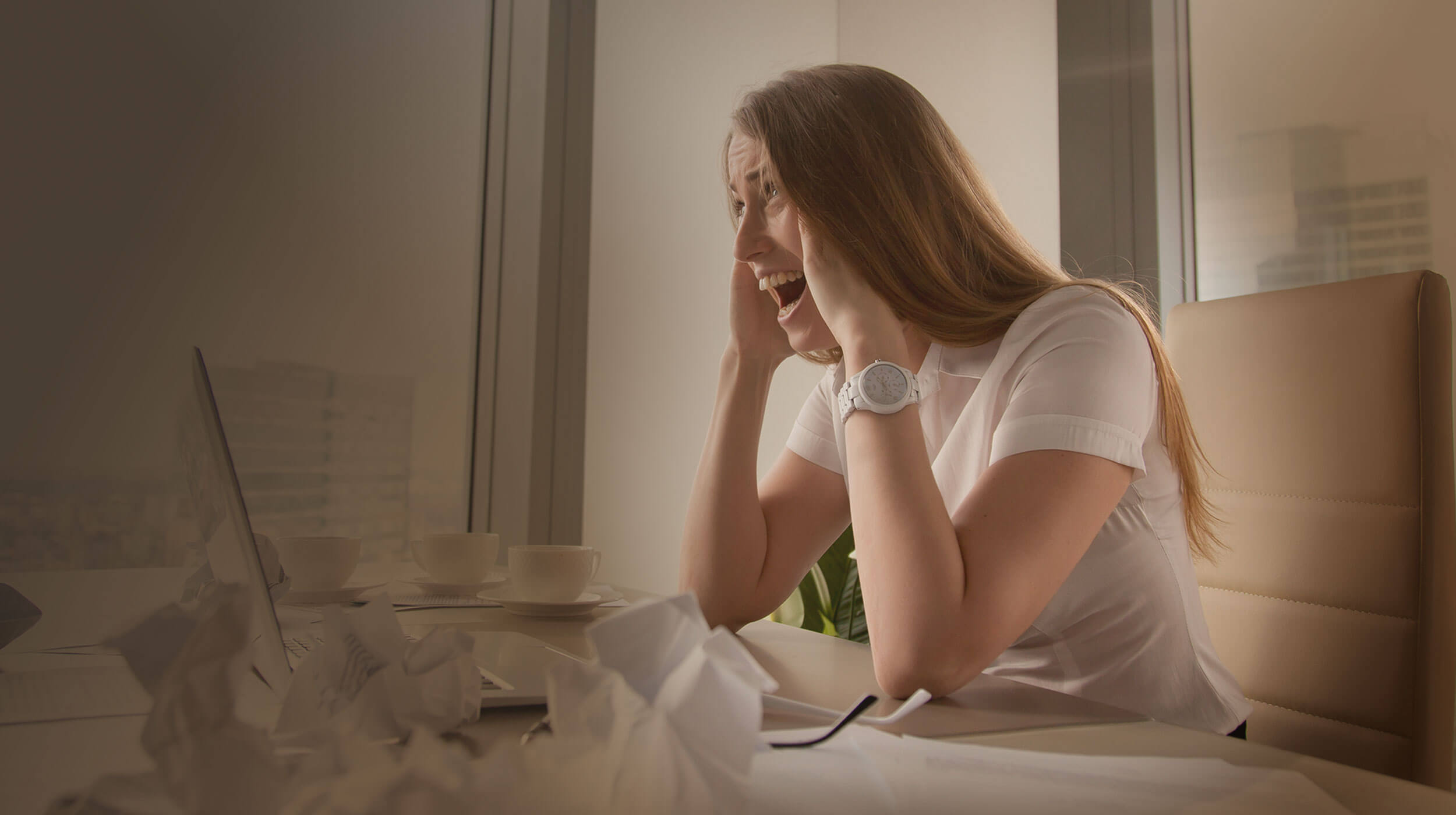 γυναίκα σε γραφείο εργασίας, απογοητευμένη και παρουσιάζοντας σημάδια burnout, με χαρτιά και άδεια φλιτζάνια καφέ γύρω της.